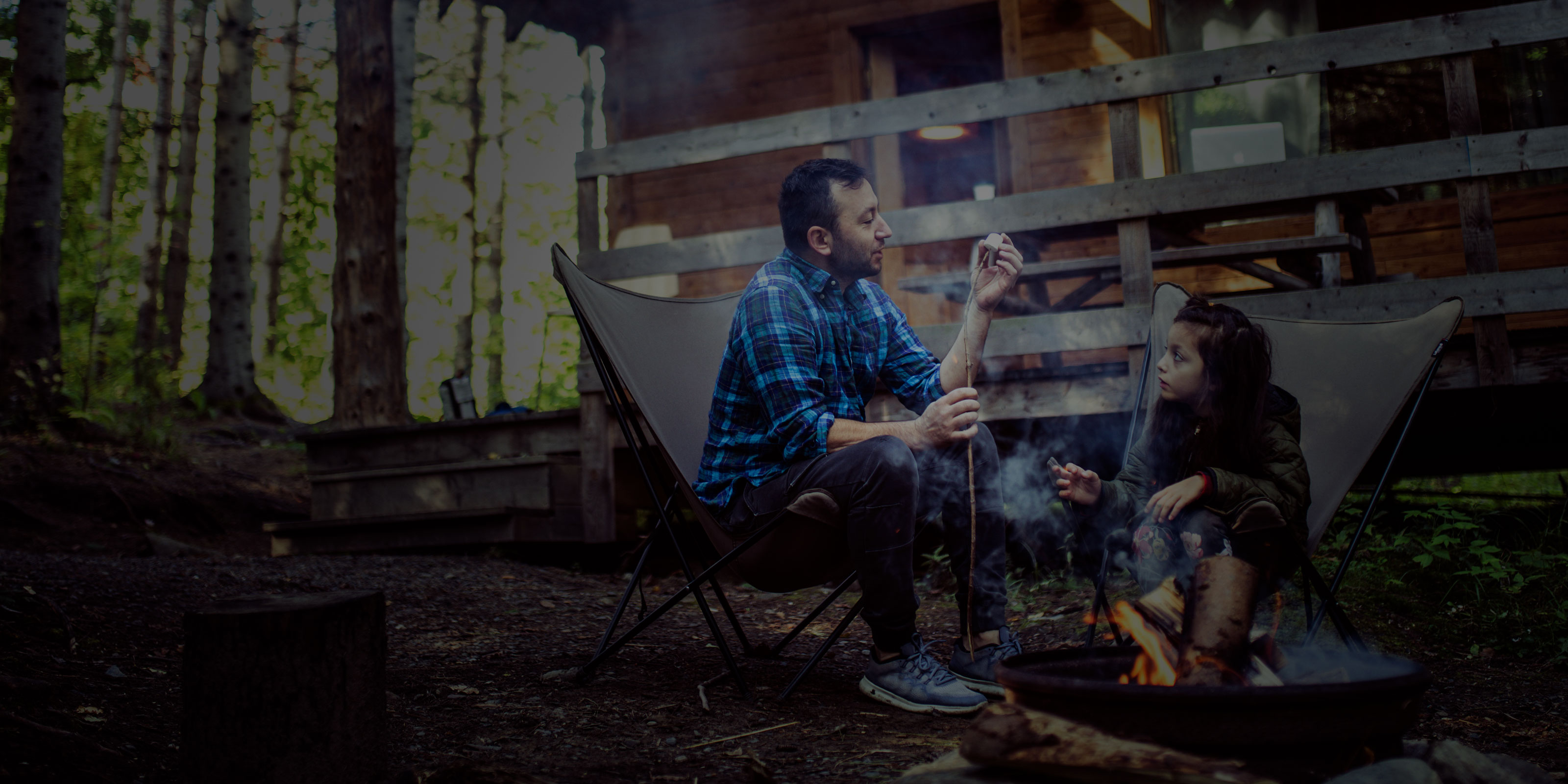 Father and daughter sitting by a lit campfire making s’mores at their cottage 